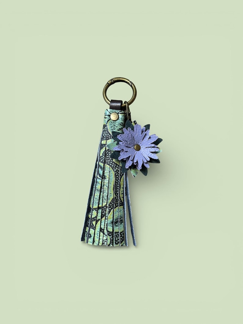 May include: A decorative keychain with a floral design. The keychain features a tassel with a green and black pattern, a metal ring, and a purple flower accent. The flower has a gold centre and is made of layered petals.