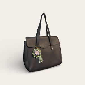 May include: A black leather tote bag with a flap closure and two top handles. A decorative floral charm with pink and green accents hangs from one handle. The bag has a structured design and a minimalist aesthetic.