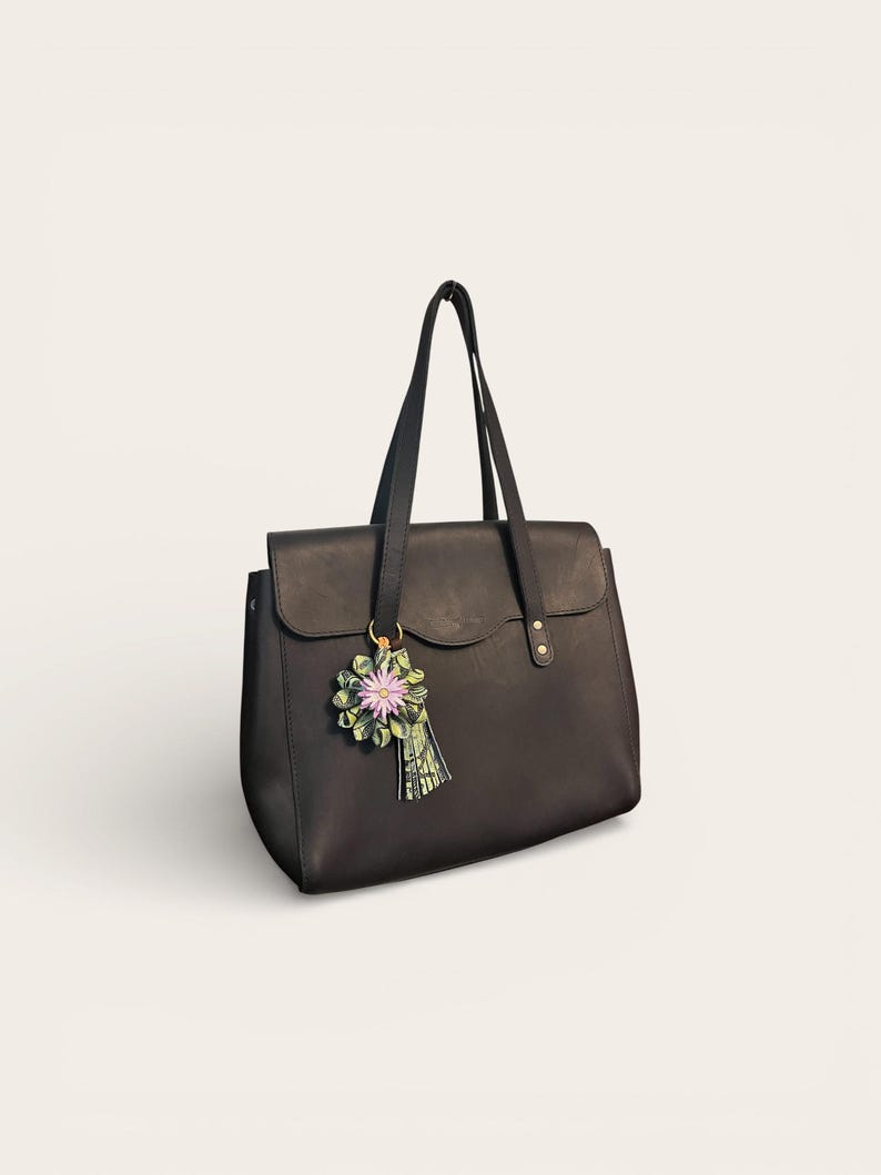 May include: A black leather tote bag with a flap closure and two top handles. A decorative floral charm with pink and green accents hangs from one handle. The bag has a structured design and a minimalist aesthetic.