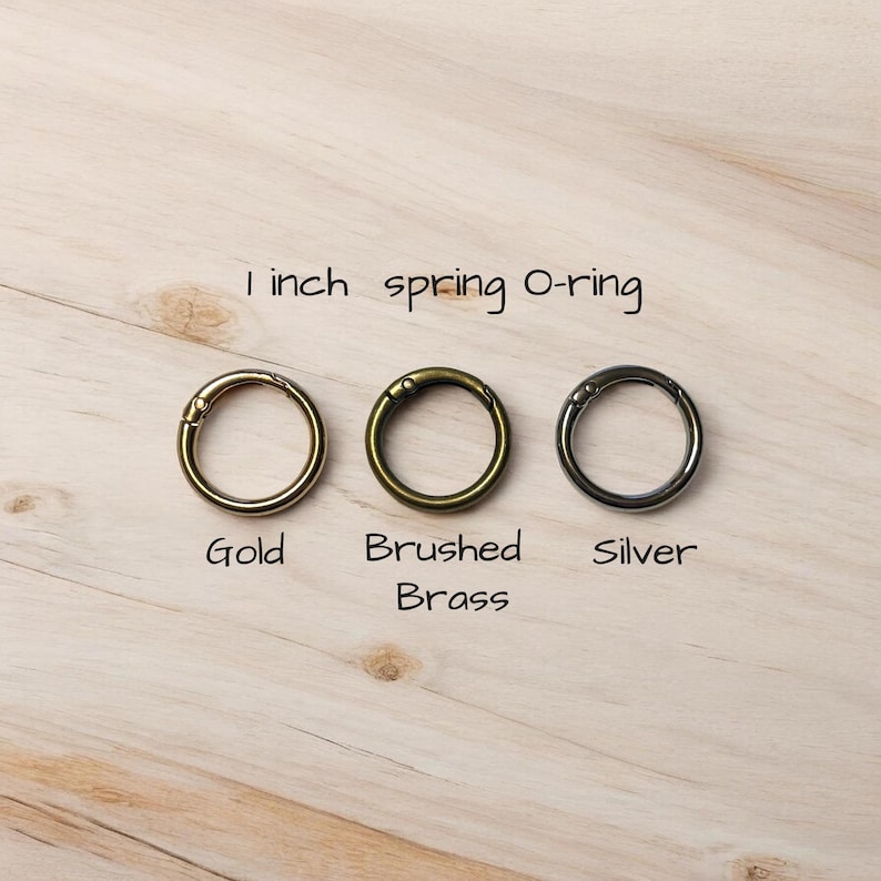May include: Three spring O-rings in gold, brushed brass, and silver finishes. The text "1 inch spring O-ring" is above the rings. The rings are approximately 2.5 cm in diameter. The gold and brass rings have a warm metallic sheen, while the silver ring has a cool tone.