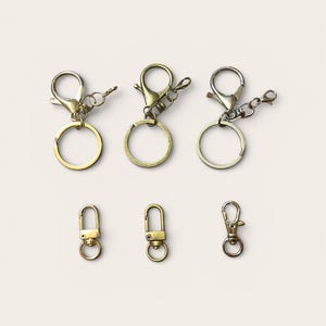 May include: An assortment of keychains and clasps in metallic finishes. The image displays three keychains with round rings and lobster claw clasps, and three smaller clasps, in gold and silver tones.