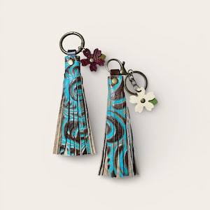 May include: Two leather keyrings with turquoise and brown swirl patterns. Each keyring has a tassel design and a decorative flower charm. The keyrings are attached to bronze-coloured metal rings.