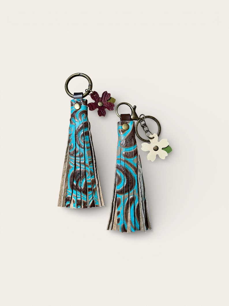 May include: Two leather keyrings with turquoise and brown swirl patterns. Each keyring has a tassel design and a decorative flower charm. The keyrings are attached to bronze-coloured metal rings.