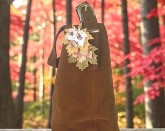 May include: A brown suede hobo bag featuring a top handle with a silver-toned buckle. A decorative charm consisting of three layered leaf-shaped cutouts in white, yellow, and olive green with floral and butterfly patterns hangs from the handle.