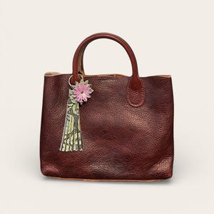 May include: A burgundy leather tote bag with a top handle. A pink flower and green tassel charm hangs from the handle. The bag has a textured surface and a simple, classic design.