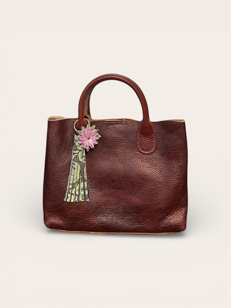 May include: A burgundy leather tote bag with a top handle. A pink flower and green tassel charm hangs from the handle. The bag has a textured surface and a simple, classic design.