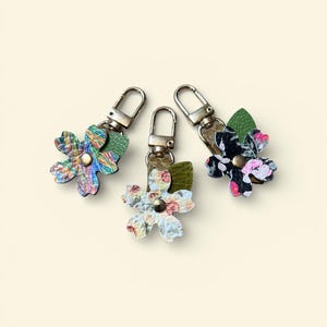 May include: Three floral keychains with silver-tone clasps. Each flower has a unique pattern: one with a colorful, iridescent design, one with a floral print, and another with a black and pink floral pattern. Each keychain has a green leaf accent.