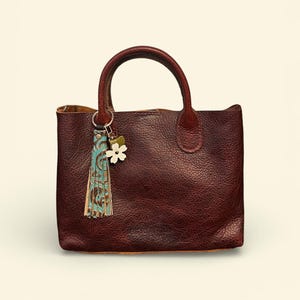 May include: A burgundy leather tote bag with a top handle. The bag has a textured surface and a small decorative tassel and flower charm attached to the handle. The tassel has blue and brown accents.