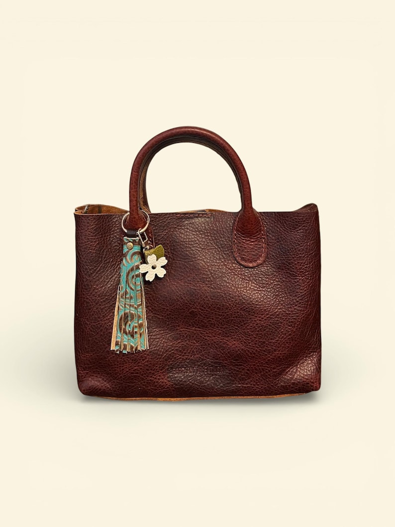 May include: A burgundy leather tote bag with a top handle. The bag has a textured surface and a small decorative tassel and flower charm attached to the handle. The tassel has blue and brown accents.