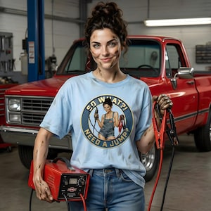 May include: A light blue t-shirt with the text "SO WHAT?! NEED A JUMP?" and a graphic of a woman holding jumper cables. The woman is holding a red battery charger and jumper cables. A red pickup truck is in the background.