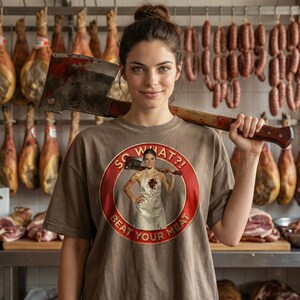 May include: A woman wearing a brown t-shirt with a graphic that says "SO WHAT?! BEAT YOUR MEAT" and holding a large meat cleaver. The background features hanging cured meats and sausages, suggesting a butcher shop setting.