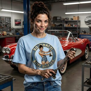 May include: A light blue t-shirt with the text "SO WHAT?! STIFF SHIFTER" and a graphic of a woman holding a gear shift. The woman is in a garage with a red classic car in the background. The person is holding a gear shift.