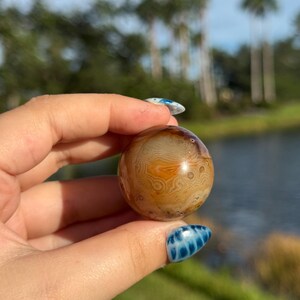 May include: A polished agate sphere with intricate patterns in shades of brown, orange, and cream. The gemstone ball is held up against a blurred background of a lake and trees.