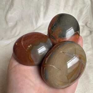 May include: Three polished, oval-shaped stones in shades of brown, gray, and red. The stones have a smooth, glossy surface and varying patterns. The stones are held in a hand against a white background.