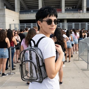 May include: Transparent backpack with black detailing. The backpack features a front pocket and mesh side pockets. A person wearing a white t-shirt and sunglasses is wearing the backpack while standing in a queue.