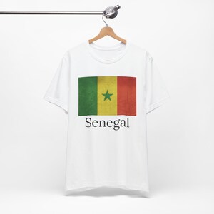 May include: White t-shirt featuring the flag of Senegal. The flag design includes green, yellow, and red vertical stripes with a green star in the center. The word "Senegal" is printed below the flag.