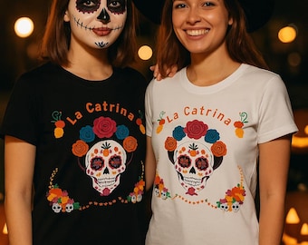 Könnte beinhalten: Ein schwarzes T-Shirt und ein weißes T-Shirt, jeweils mit einem Grafikdruck eines stilisierten Zuckerschädels, der mit bunten Blumen und dem Text La Catrina verziert ist. Das Schädeldesign enthält komplizierte Muster in roter, oranger, blauer und schwarzer Tinte.