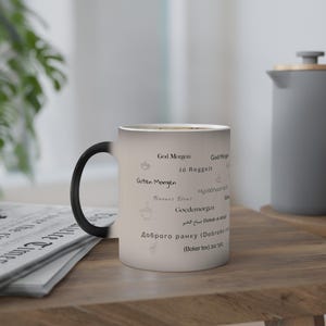 以下が含まれることがあります： 黒い持ち手が付いた白いセラミックコーヒーマグ。マグカップには、「God Morgon」、「Guten Morgen」、「Buenos Dias」など、さまざまな言語の挨拶がプリントされています。木製のテーブルの上に置かれ、新聞と灰色のケトルが背景にあります。