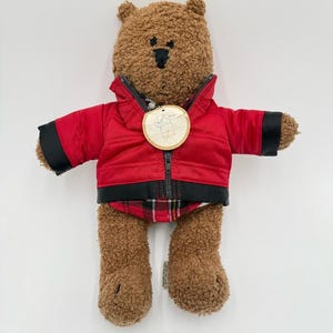 May include: A brown teddy bear with textured fur, wearing a red jacket with black trim and a red and black plaid shirt underneath. A round wooden tag with a line drawing of a bear is attached to the jacket zipper. The bear has black stitched eyes.