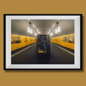 May include: A framed photograph of a subway station with a vending machine in the centre. Two yellow trains are blurred on either side of the platform. Overhead, there are several hanging lights.