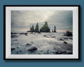 May include: A framed landscape photograph showing sea stacks in the ocean under a cloudy sky. The sea stacks are tall, rocky formations with green vegetation. The water is blurred, milky white, and the foreground rocks are dark.