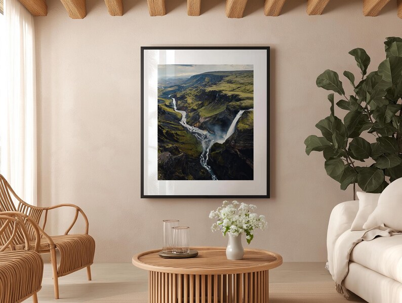 Pu&ograve; includere: Fotografia incorniciata di una cascata in un canyon verde e marrone. L'immagine &egrave; esposta in un soggiorno con un tavolino in legno, sedie in vimini e un divano bianco. La stanza ha una combinazione di colori neutri.
