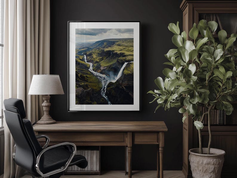 Pu&ograve; includere: Fotografia incorniciata di un paesaggio di cascata in una valle verde. L'opera d'arte &egrave; esposta su una parete scura sopra una scrivania in legno con una lampada e una sedia da ufficio nera. Una pianta in vaso &egrave; a destra.