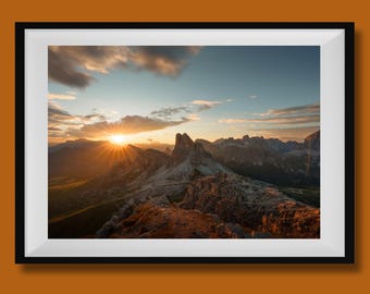May include: A framed landscape photograph captures a mountain range at sunset. The sun's rays burst through the clouds, illuminating the peaks and valleys. The image is framed in a black border with a white mat.