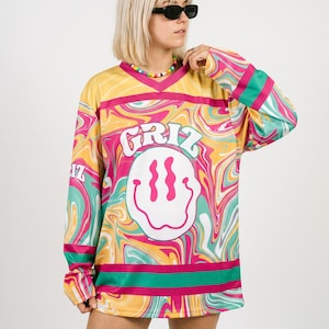 May include: A long-sleeve V-neck jersey featuring a vibrant, multi-colored psychedelic swirl pattern in yellow, pink, and teal. The front displays the text GRIZ above a large white smiley face graphic with pink accents and a pink border.