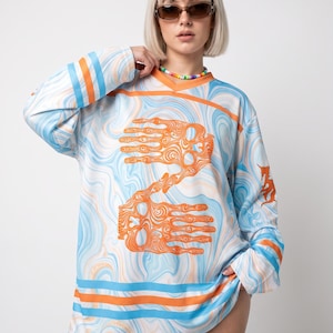 May include: A long-sleeve V-neck jersey featuring a light blue and white marbled swirl pattern with orange accents. The front displays two large orange hand graphics with intricate line details, and the sleeves have horizontal orange and blue stripes.
