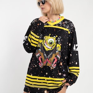 May include: A black long-sleeve jersey featuring a galaxy print with yellow stripes on the sleeves and hem. The front displays a graphic of a yellow one-eyed monster above two snarling brown dogs with chains. The number 42 is printed on the sleeve.