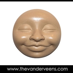 Pode incluir: Uma escultura de cerâmica de um rosto sorridente com os olhos fechados. O rosto é de cor castanho claro e tem uma textura lisa e arredondada.