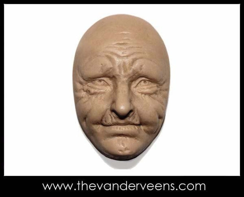 Puede incluir: Una escultura realista y detallada de un rostro humano, esculpida en un color marr&oacute;n claro. El rostro tiene arrugas y l&iacute;neas profundas, lo que sugiere una persona mayor. La escultura tiene forma ovalada y parece estar hecha de arcilla o yeso.