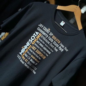 May include: Black t-shirt with a white graphic design featuring a list of Minnesota state parks and landmarks. The text includes "Minnesota", "North Shore", "Lake Superior", "Jay Cooke SP", "Duluth Canal Park", "Gooseberry Falls State Park", "Tettegouche State Park", "Palisade Head", "Black Sand Beach", "Split Rock Lighthouse", "Temperance River State Park", "Cascade River State Park", "Gunflint Grand Marais", "Judge C.R. Magney SP", "Hwy 61", "Devil's Kettle", and "Grand Portage State Park".