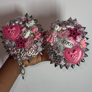 May include: Two heart-shaped accessories, heavily embellished with pink, silver, and clear rhinestones, charms, and spikes. The word "Barbie" is written in cursive. The accessories feature pink hearts, stars, and cartoon figures.
