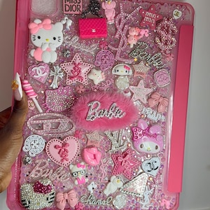 Könnte beinhalten: Eine rosa Tablet-Hülle, die mit einer dichten Collage aus Anhängern verziert ist, darunter Hello Kitty-Figuren, Barbie-Logos, Marken-Embleme wie Prada, Chanel und Dior, rosa Sterne, Schleifen, Herzen und funkelnde Strasssteine auf der Oberfläche.
