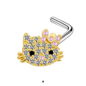 Pode incluir: Um piercing de nariz em forma de L com uma cabeça de gato dourada adornada com cristais brancos transparentes, dois olhos de cristal preto e um grupo de cristais cor-de-rosa formando um laço na lateral, fixado num poste de metal prateado.