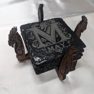 Puede incluir: Un juego de posavasos cuadrados de pizarra con un soporte de madera decorativo. Cada posavasos es negro con un diseño grabado en gris con la letra "M" y la palabra "MAY". El soporte de madera tiene detalles en forma de dragón.