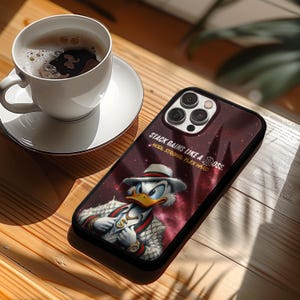 May include: A smartphone case featuring a cartoon duck in a suit and hat, with the text "STACK GAINS LIKE A BOSS. HODL STRONG. FLEX HARD." The case has a dark red and purple background with a starry design. A cup of coffee sits on a saucer in the background.