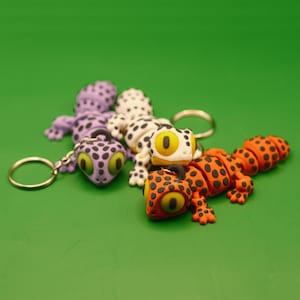 Könnte beinhalten: Drei farbige Gecko-Schlüsselanhänger mit schwarzen Punkten. Die Geckos sind lila, weiß und orange mit schwarzen Punkten. Die Schlüsselanhänger sind an silbernen Schlüsselringen befestigt.