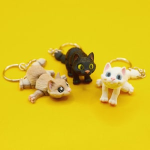 Peut inclure: Trois porte-clés en forme de chat, un noir, un blanc et un brun, avec un design ludique. Chaque porte-clés représente un chat avec de grands yeux et une expression mignonne.