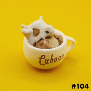 May include: A white ceramic mug with the word "Cubone" in brown script. Inside the mug is a brown and white figurine of a Cubone character. The background is a solid yellow.