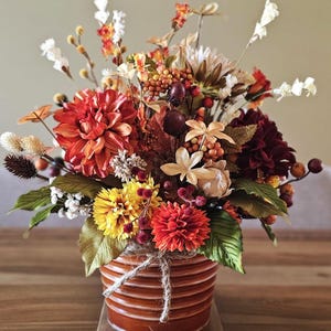May include: A vibrant floral arrangement in a brown ceramic pot. The bouquet features orange, red, yellow, and white flowers, with green leaves and berries. The pot has a rope tied around it. The arrangement is on a wooden surface.