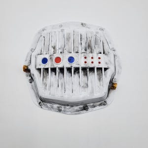 May include: A white differential cover with a distressed black finish, featuring vertical cooling fins, three circular insets in blue and red, and a rectangular section with six small red dots. Two brass-coloured fittings are on the sides.