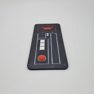 May include: A rectangular black control panel featuring a textured surface with a red circular button, three white square buttons, and a red rectangular indicator light. The panel includes silver-colored accents and small circular screw-like details.