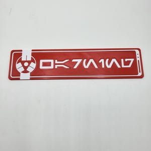 Könnte beinhalten: Ein rechteckiges rotes Schild mit weißem Rand und weißem Text, der OK7W1W7 lautet. Auf der linken Seite befindet sich ein weißes kreisförmiges Emblem mit einem stilisierten Strahlensymbol. Das Objekt hat eine flache, glatte Oberfläche.