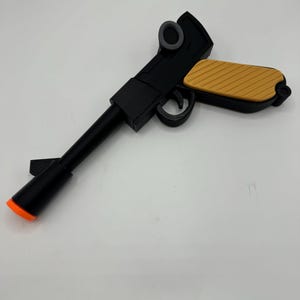 May include: A black toy pistol featuring a long barrel with an orange tip, a trigger guard, and a textured mustard yellow grip. The design includes a circular gray detail on the upper frame, presenting a sleek, angular construction throughout.