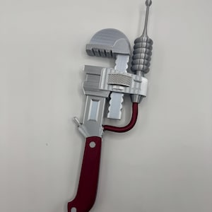 May include: A silver-colored pipe wrench prop featuring a maroon handle and a maroon curved accent piece. The tool includes a silver antenna-like attachment with a ribbed cylindrical section and a small sphere at the top, all set against a plain background.