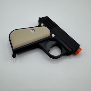 May include: A black toy pistol featuring a matte finish, a cream-colored grip panel secured with two silver-toned screws, a trigger guard, and a bright orange safety tip at the end of the barrel. The design includes textured slide serrations.