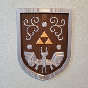 May include: A shield-shaped wall plaque featuring a brown base with a silver border. The centre displays a gold-coloured Triforce symbol above a silver-coloured winged creature emblem, flanked by silver scrollwork and circular accents on the brown surface.
